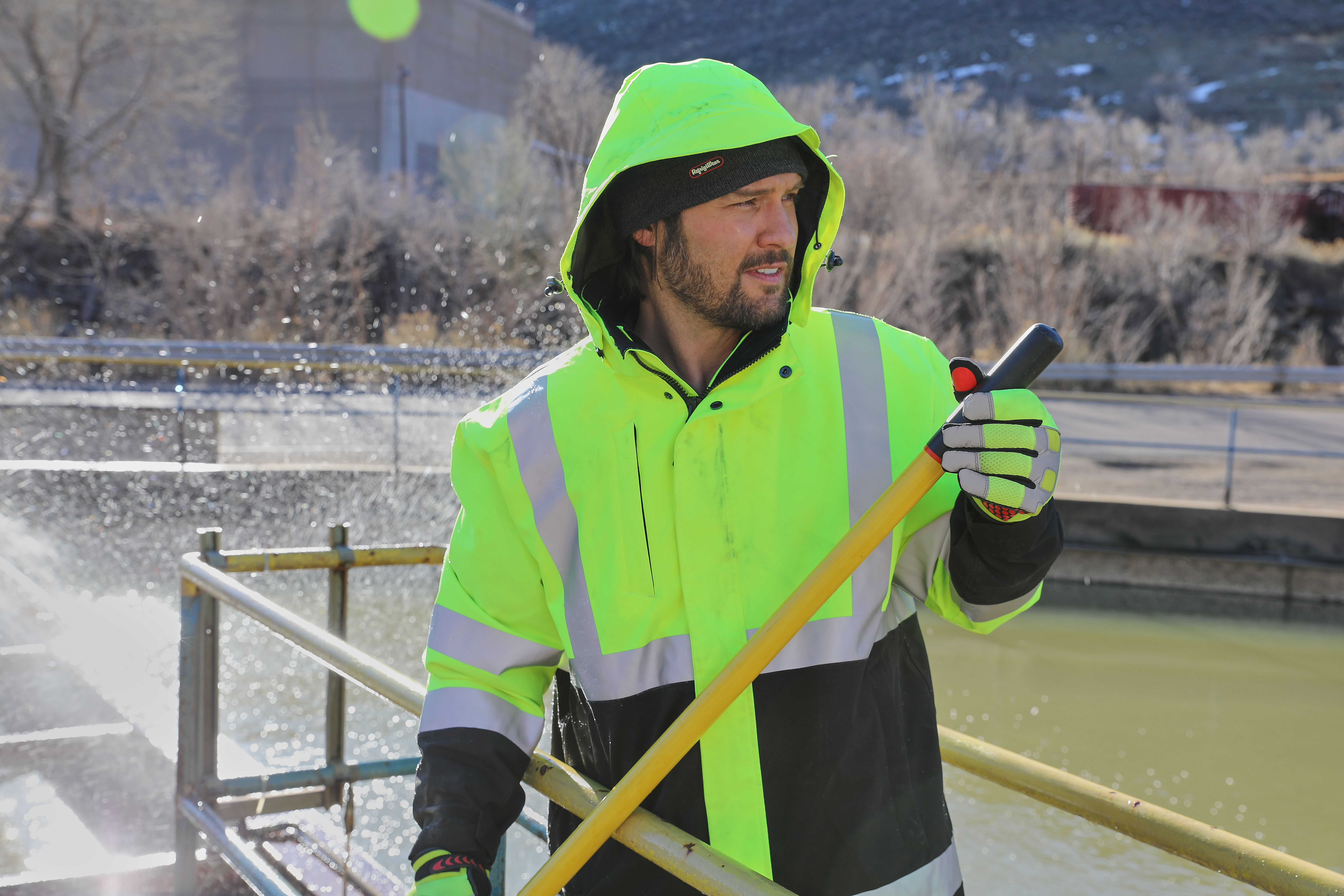 Worker in neon yellow safety jacket and hood holding shovel by waterfront