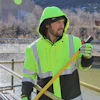 Worker in neon yellow safety jacket and hood holding shovel by waterfront