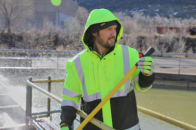 Worker in neon yellow safety jacket and hood holding shovel by waterfront