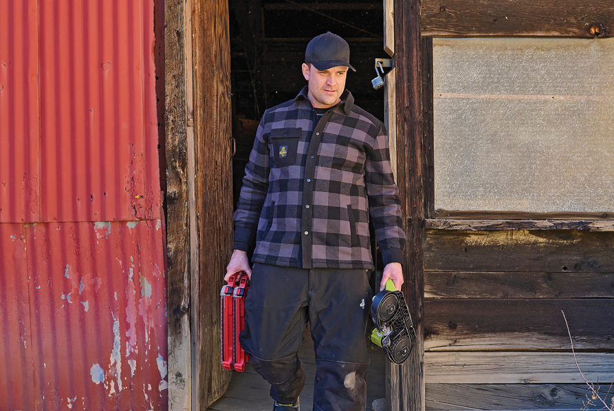 Man in blue plaid shirt holding tools stands in doorway of weathered wooden building with red metal siding