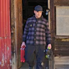 Man in blue plaid shirt holding tools stands in doorway of weathered wooden building with red metal siding