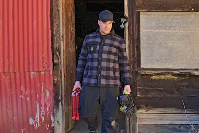 Man in blue plaid shirt holding tools stands in doorway of weathered wooden building with red metal siding