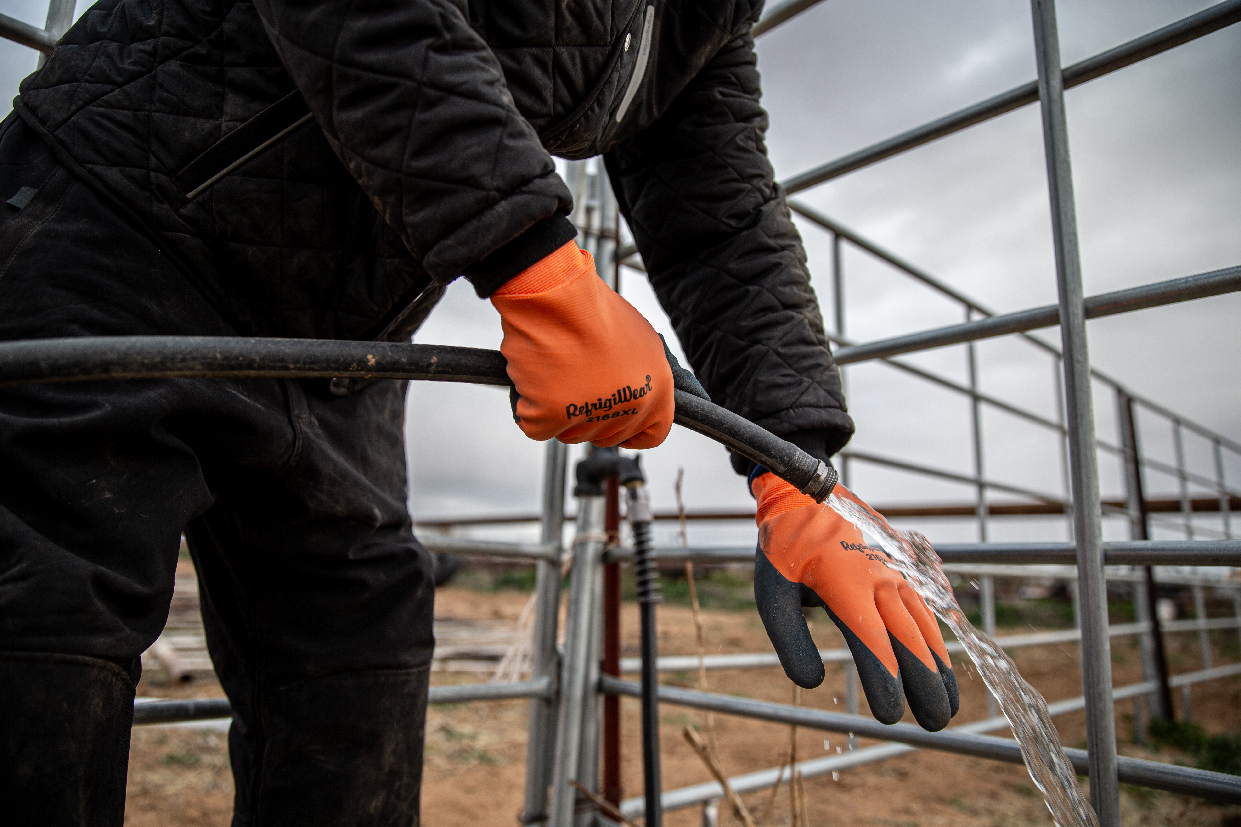 Worker in black jacket wearing orange Mechanix work gloves while handling metal railing at industrial facility