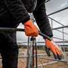 Worker in black jacket wearing orange Mechanix work gloves while handling metal railing at industrial facility