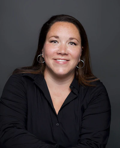 Professional headshot of a woman with long dark hair wearing a black button-up shirt and silver hoop earrings, smiling against a gray background