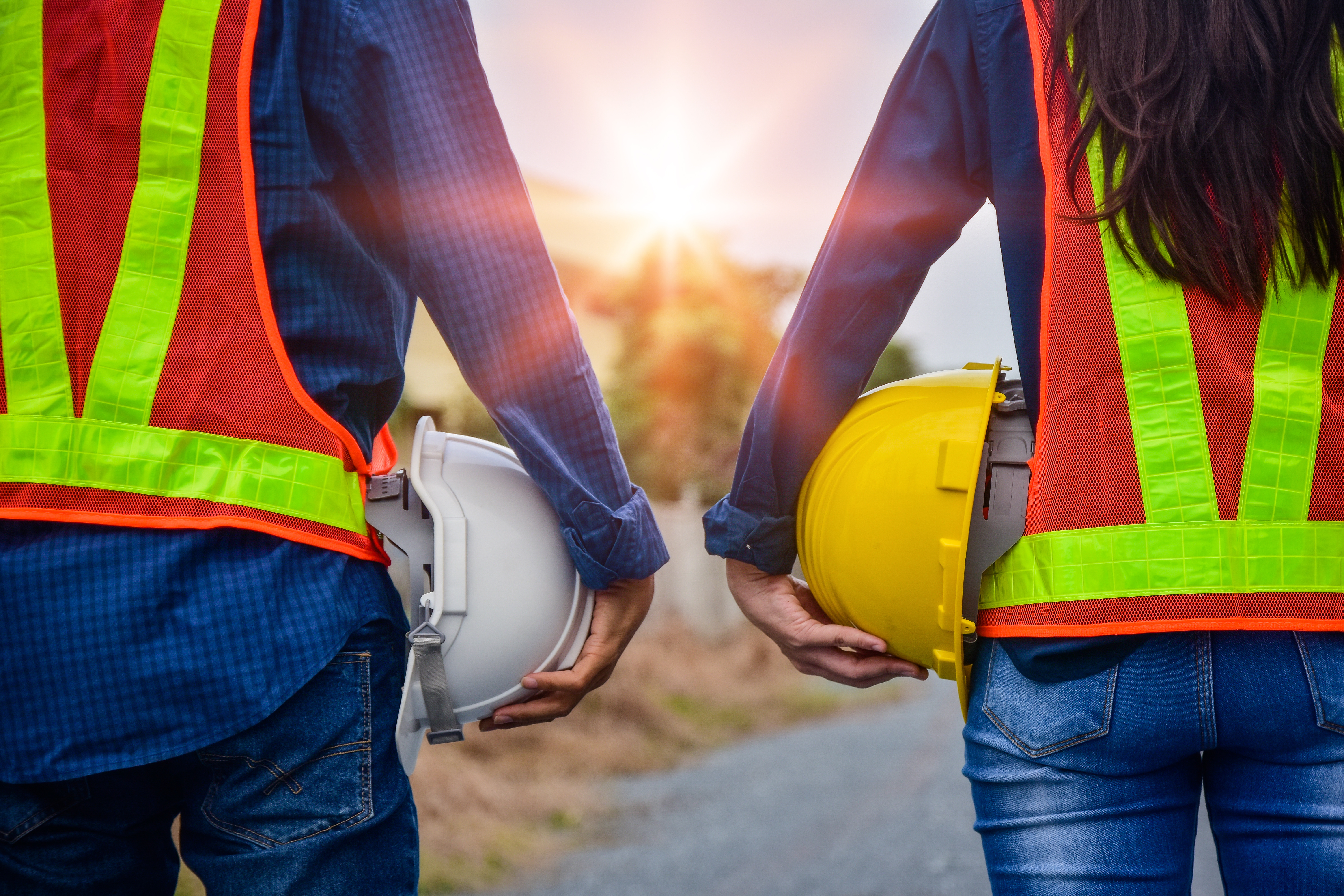 Two construction workers in safety vests holding white and yellow hard hats against a sunset backdrop