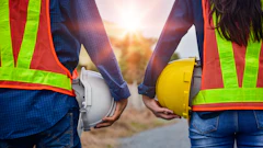 Two construction workers in safety vests holding white and yellow hard hats against a sunset backdrop
