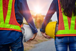Two construction workers in safety vests holding white and yellow hard hats against a sunset backdrop