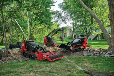 Red and black tracked mulching equipment and excavators clearing land beneath tall trees in a residential yard.