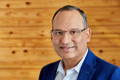 Professional headshot of a man wearing glasses and blue blazer against wooden background