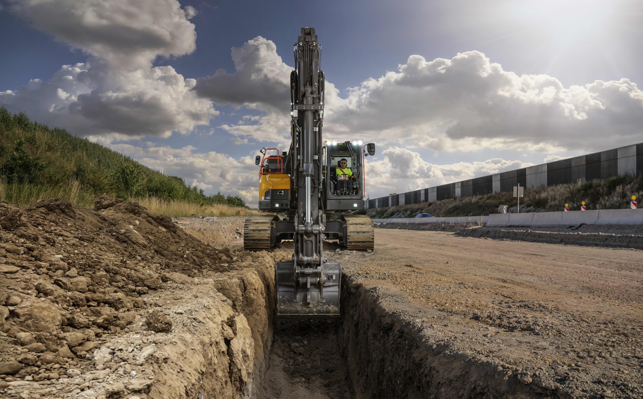 Excavator digging trench at construction site with yellow truck and industrial buildings in background under cloudy sky