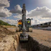 Excavator digging trench at construction site with yellow truck and industrial buildings in background under cloudy sky