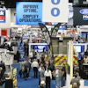 Busy trade show floor with multiple vendor booths, overhead signage, and crowds of attendees at an industrial or automotive exhibition.
