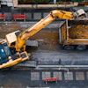 Yellow excavator loading construction debris into truck on urban street with benches and buildings