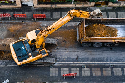 Yellow excavator loading construction debris into truck on urban street with benches and buildings