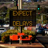 Digital traffic sign displaying 'EXPECT DELAYS' in amber text above a parking lot with vehicles and landscaping.