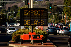 Digital traffic sign displaying 'EXPECT DELAYS' in amber text above a parking lot with vehicles and landscaping.