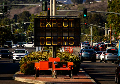 Digital traffic sign displaying 'EXPECT DELAYS' in amber text above a parking lot with vehicles and landscaping.