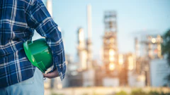 Construction worker in blue plaid shirt holding green hard hat with industrial refinery in background