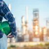 Construction worker in blue plaid shirt holding green hard hat with industrial refinery in background