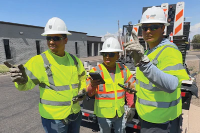 Three construction workers in neon safety vests and white hard hats pose at a job site with equipment in background