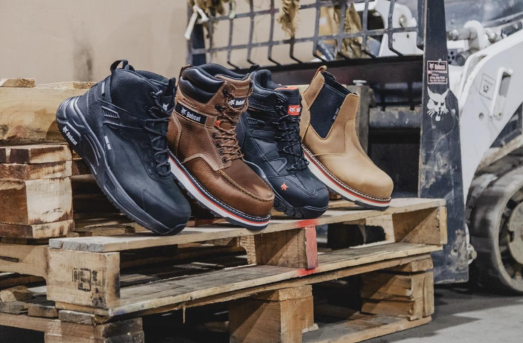 Four work boots in navy, brown, and tan displayed on wooden pallet in industrial setting