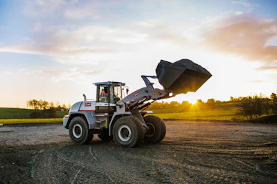 Blue and white wheel loader with raised bucket on rural road at sunset with green fields in background