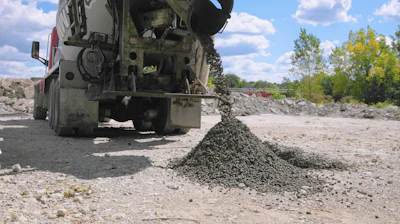 Concrete mixer truck discharging aggregate at construction site with stockpiles and trees