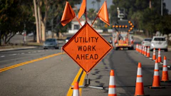 Orange diamond-shaped construction warning sign reading 'UTILITY WORK AHEAD' on a street with traffic cones and equipment