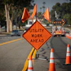 Orange diamond-shaped construction warning sign reading 'UTILITY WORK AHEAD' on a street with traffic cones and equipment