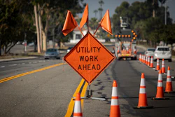 Orange diamond-shaped construction warning sign reading 'UTILITY WORK AHEAD' on a street with traffic cones and equipment