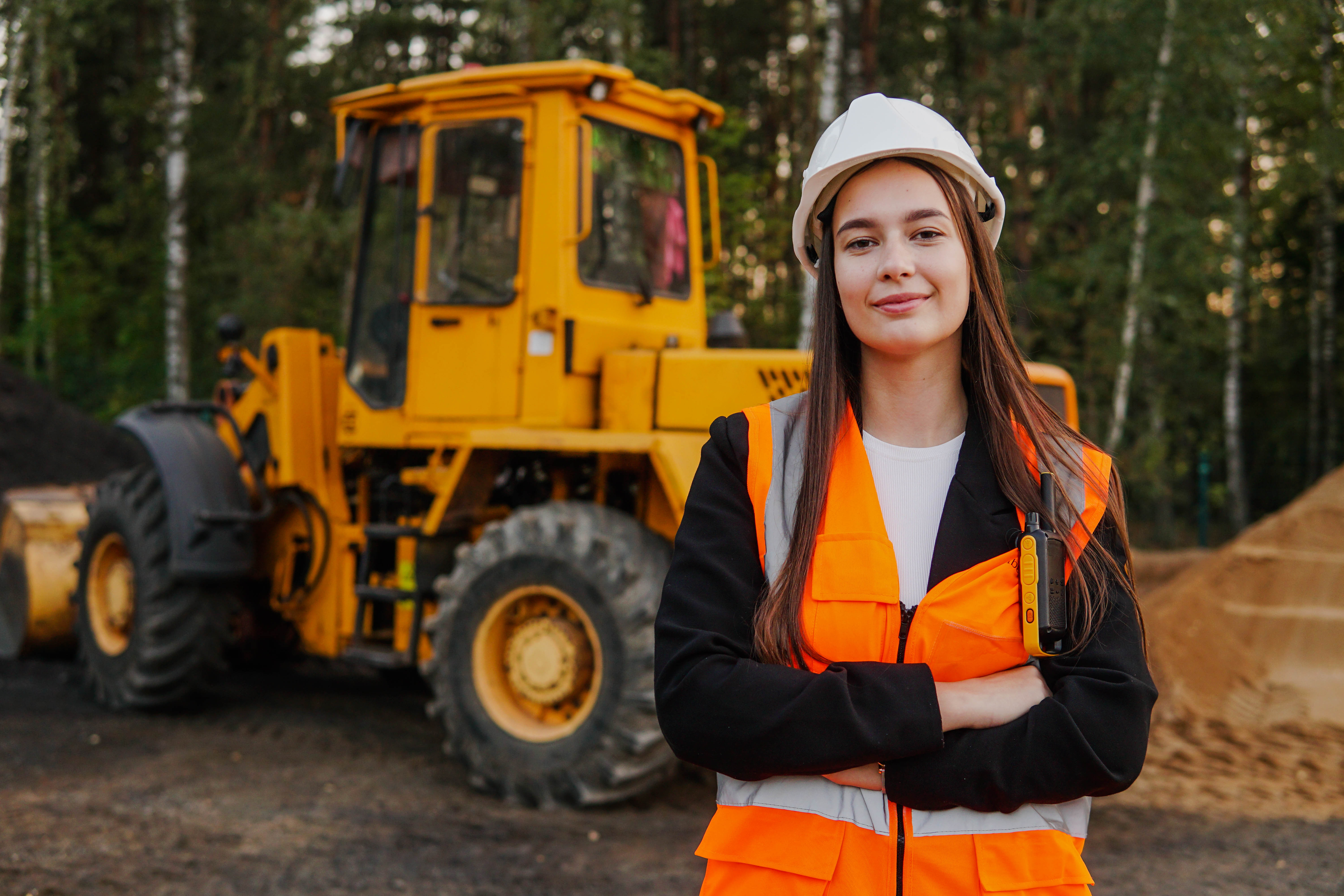 Female construction worker in safety gear smiling with yellow wheel loader in forest background