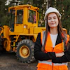 Female construction worker in safety gear smiling with yellow wheel loader in forest background
