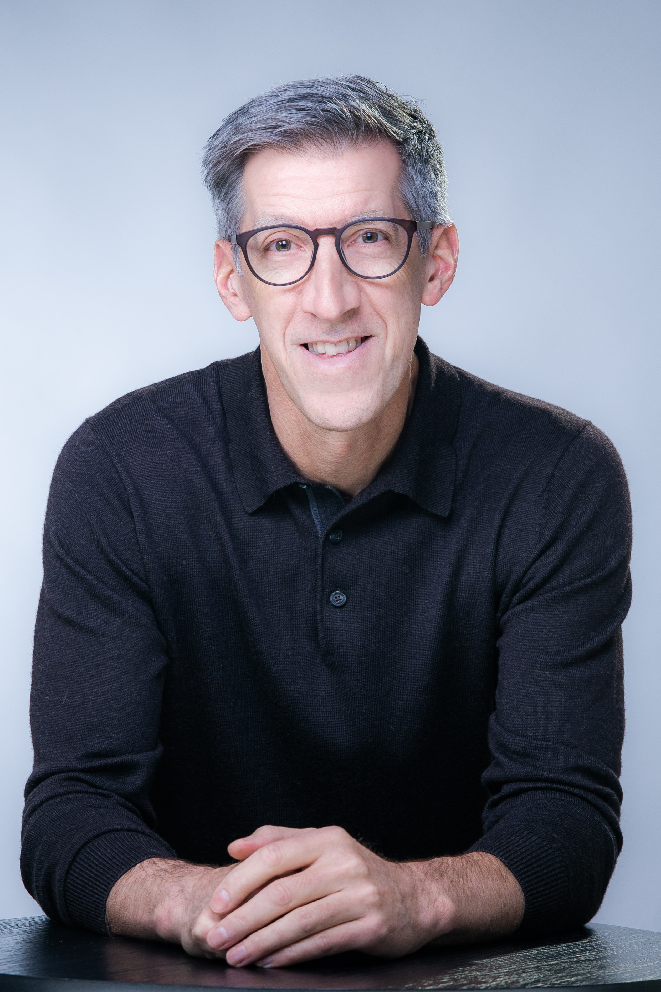 Professional headshot of a smiling man wearing glasses and a dark polo shirt against a light blue background Headshot
