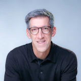 Professional headshot of a smiling man wearing glasses and a dark polo shirt against a light blue background