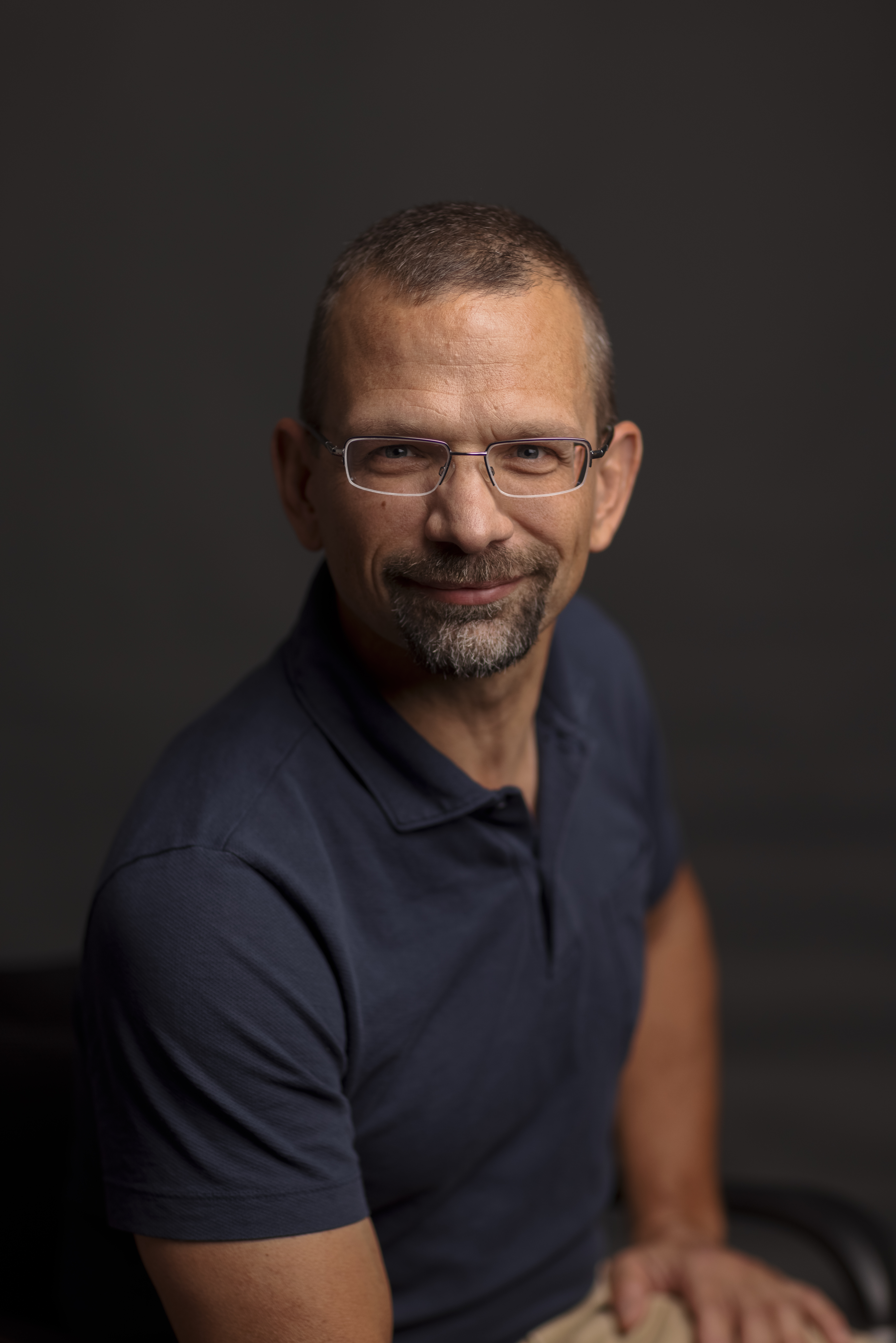 Professional headshot of a man wearing glasses and dark polo shirt against neutral background Headshot