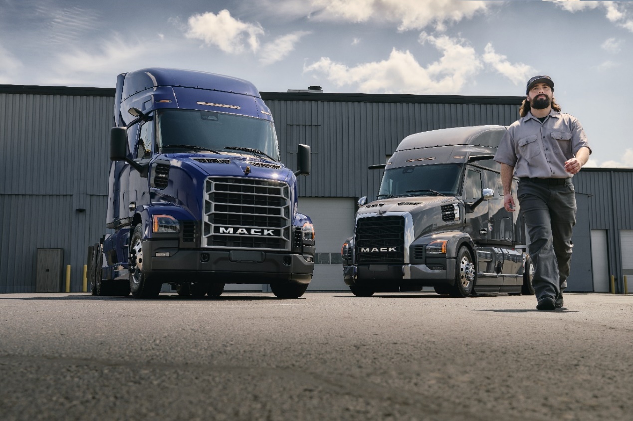 Two Mack trucks parked at an industrial facility with a worker standing between them under a cloudy sky