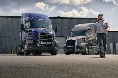 Two Mack trucks parked at an industrial facility with a worker standing between them under a cloudy sky