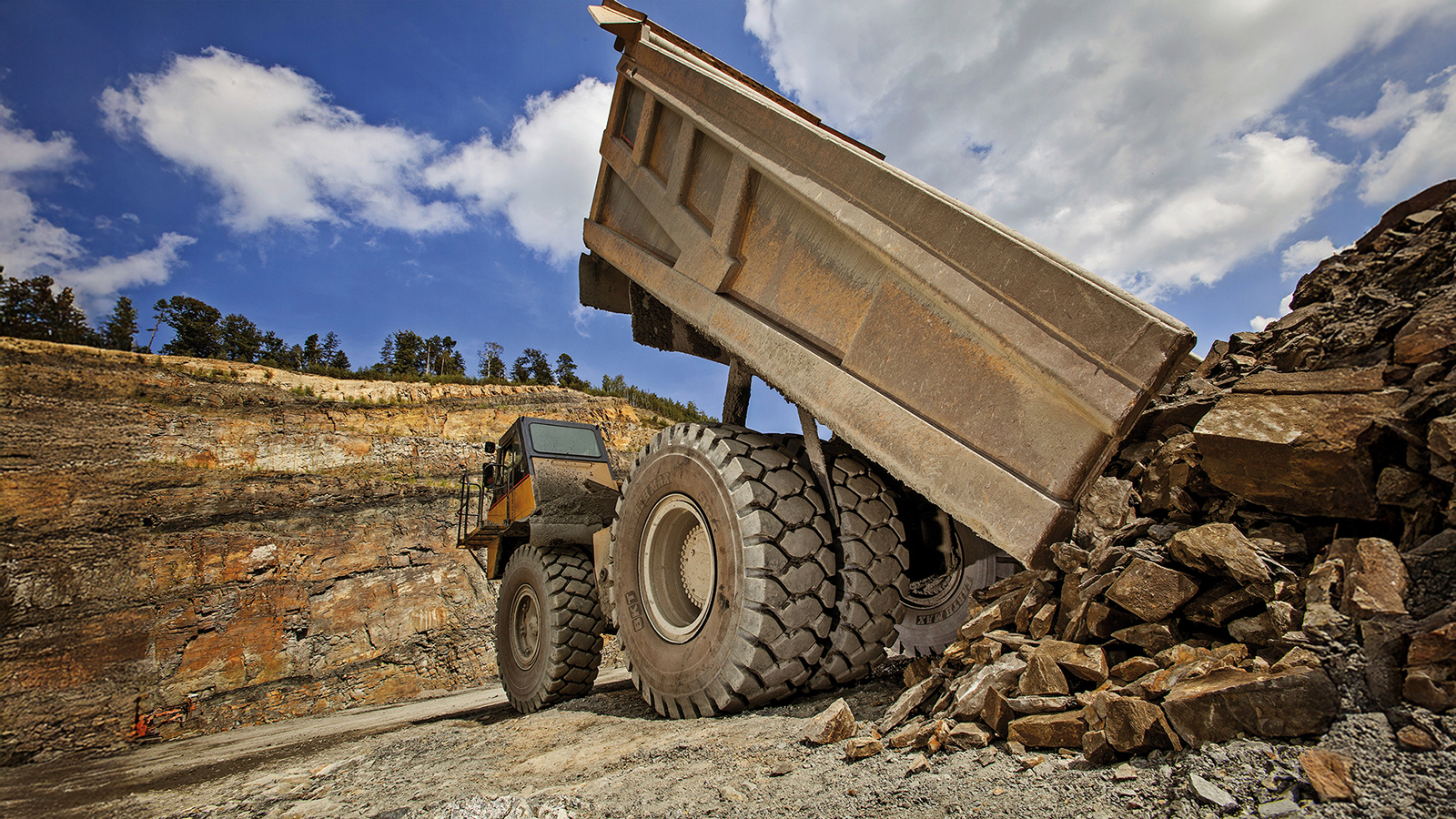 Dump truck unloading rocks and gravel at quarry with blue sky and trees in background