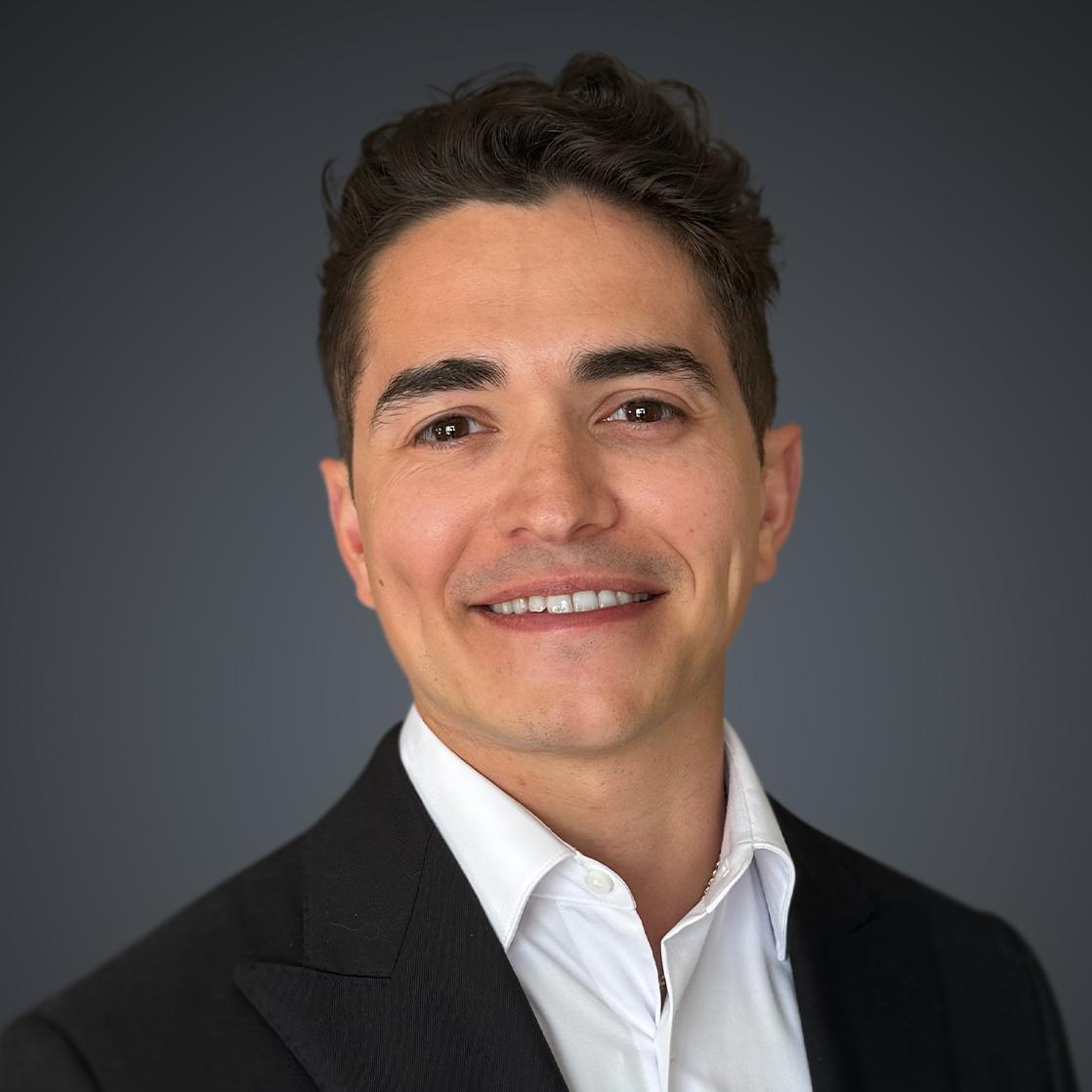 Professional headshot of a smiling man in black blazer and white shirt against gray background Headshot