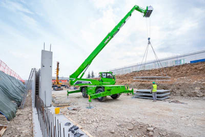 Green MERLO telescopic handler lifting construction materials at a building site with workers and equipment visible