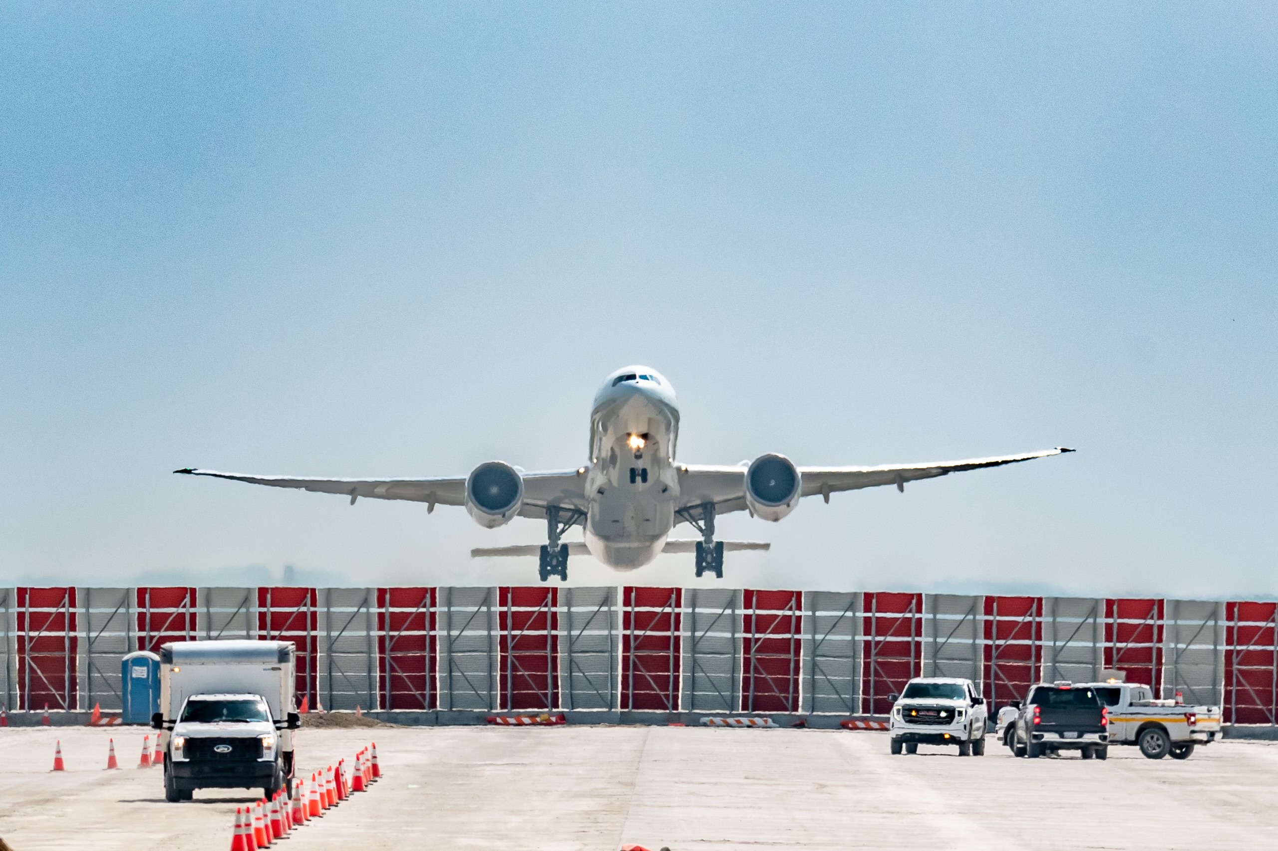 Commercial aircraft approaching airport terminal with ground support vehicles and red hangar doors visible under clear sky