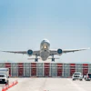 Commercial aircraft approaching airport terminal with ground support vehicles and red hangar doors visible under clear sky