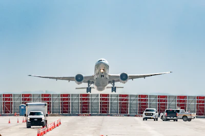 Commercial aircraft approaching airport terminal with ground support vehicles and red hangar doors visible under clear sky