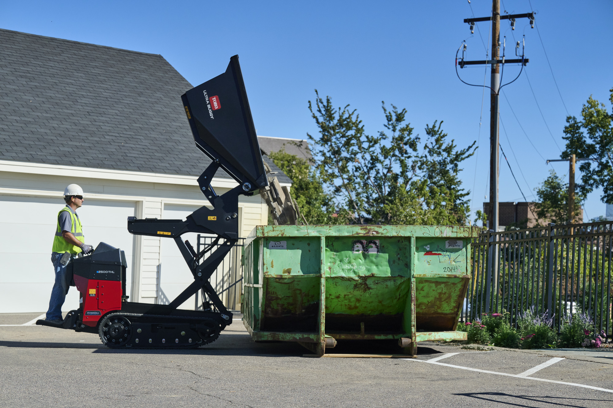 Worker operating green dumpster with red compactor truck in residential driveway on sunny day