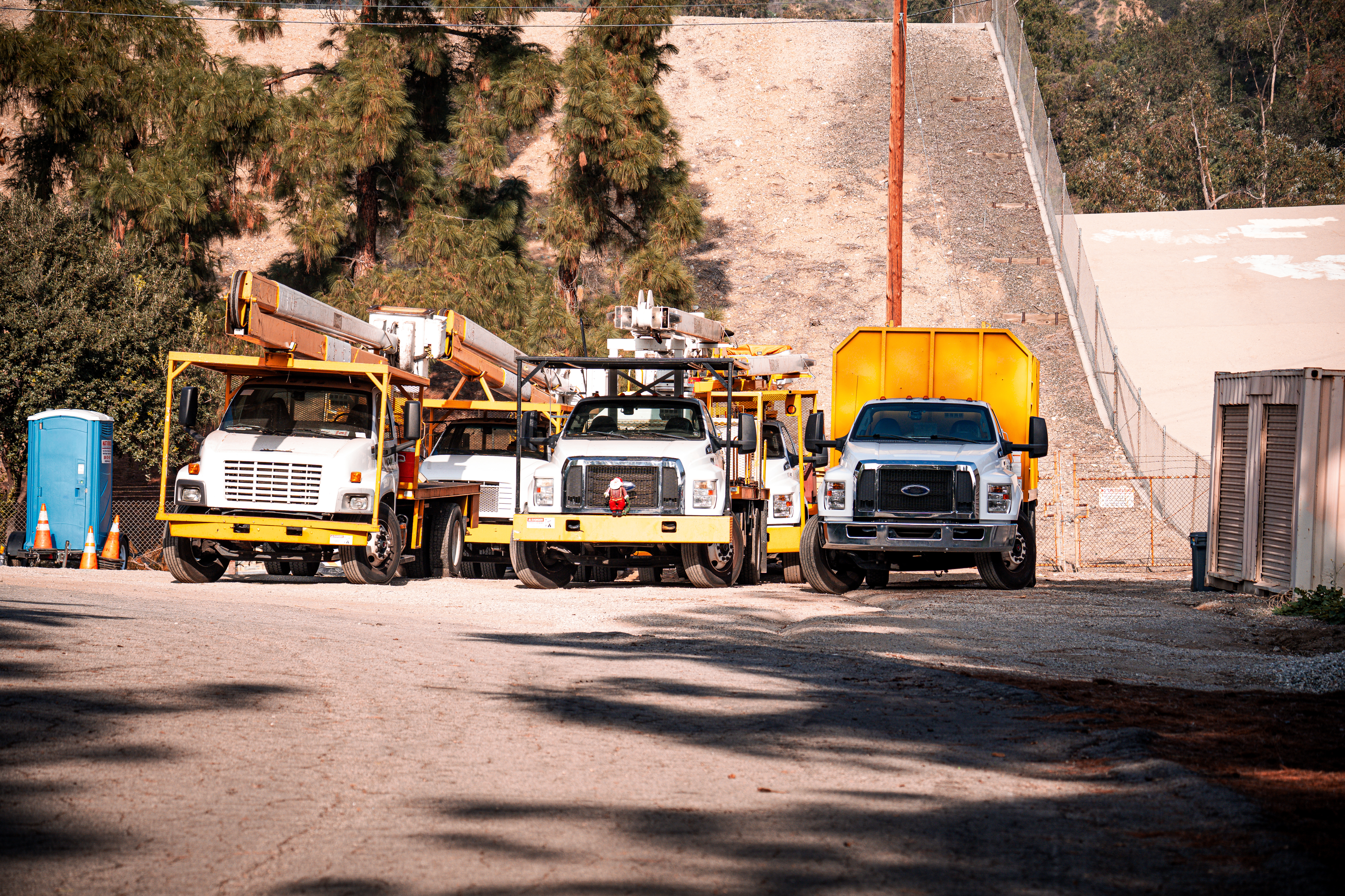 Construction utility trucks and equipment parked in a yard with trees and hillside in background