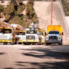 Construction utility trucks and equipment parked in a yard with trees and hillside in background