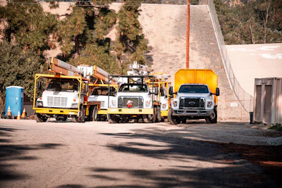 Construction utility trucks and equipment parked in a yard with trees and hillside in background