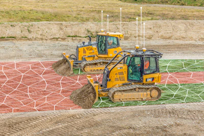 Two yellow Caterpillar bulldozers working on a sports field with red and green netting visible in the background.