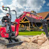 Red mini excavator with hydraulic arm operating on gravel near a rustic lodge building under blue sky